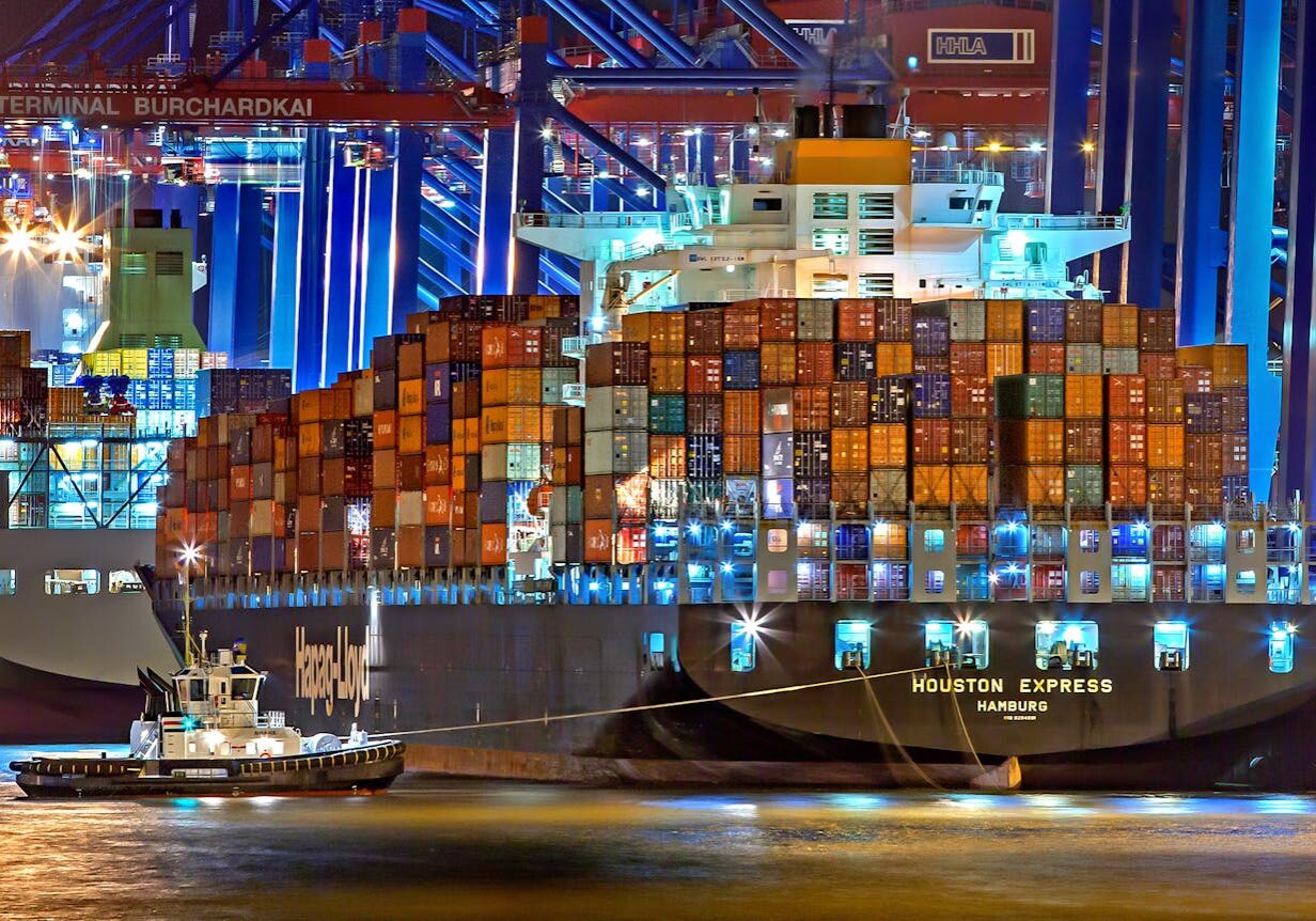 A brightly lit cargo ship at Hamburg harbor with stacked containers and a tugboat.